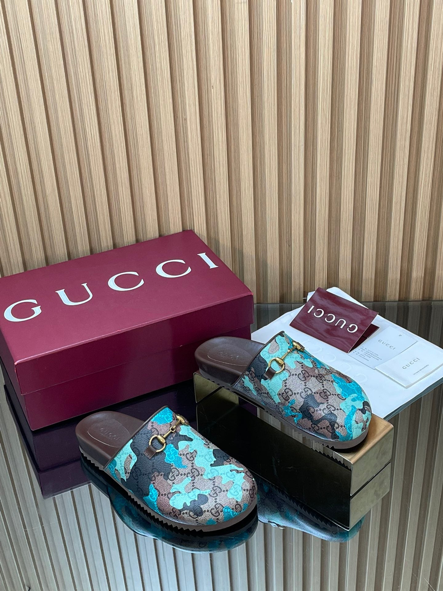 Gucci slide sandals pair, teal GG-pattern fabric strap and gold buckle on dark soles.