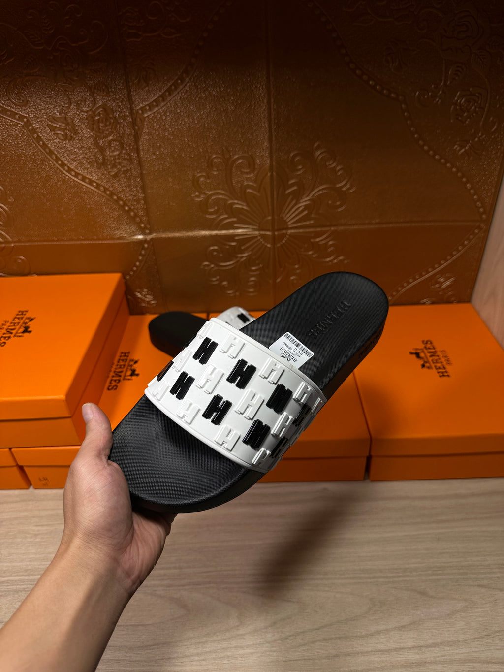 Hand holding a black slide sandal with white patterned strap and black footbed