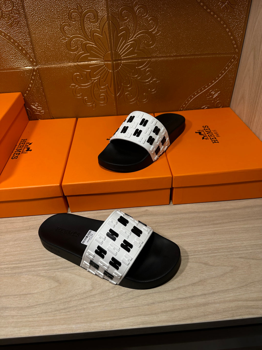 One black slide sandal with white patterned strap shown on display boxes
