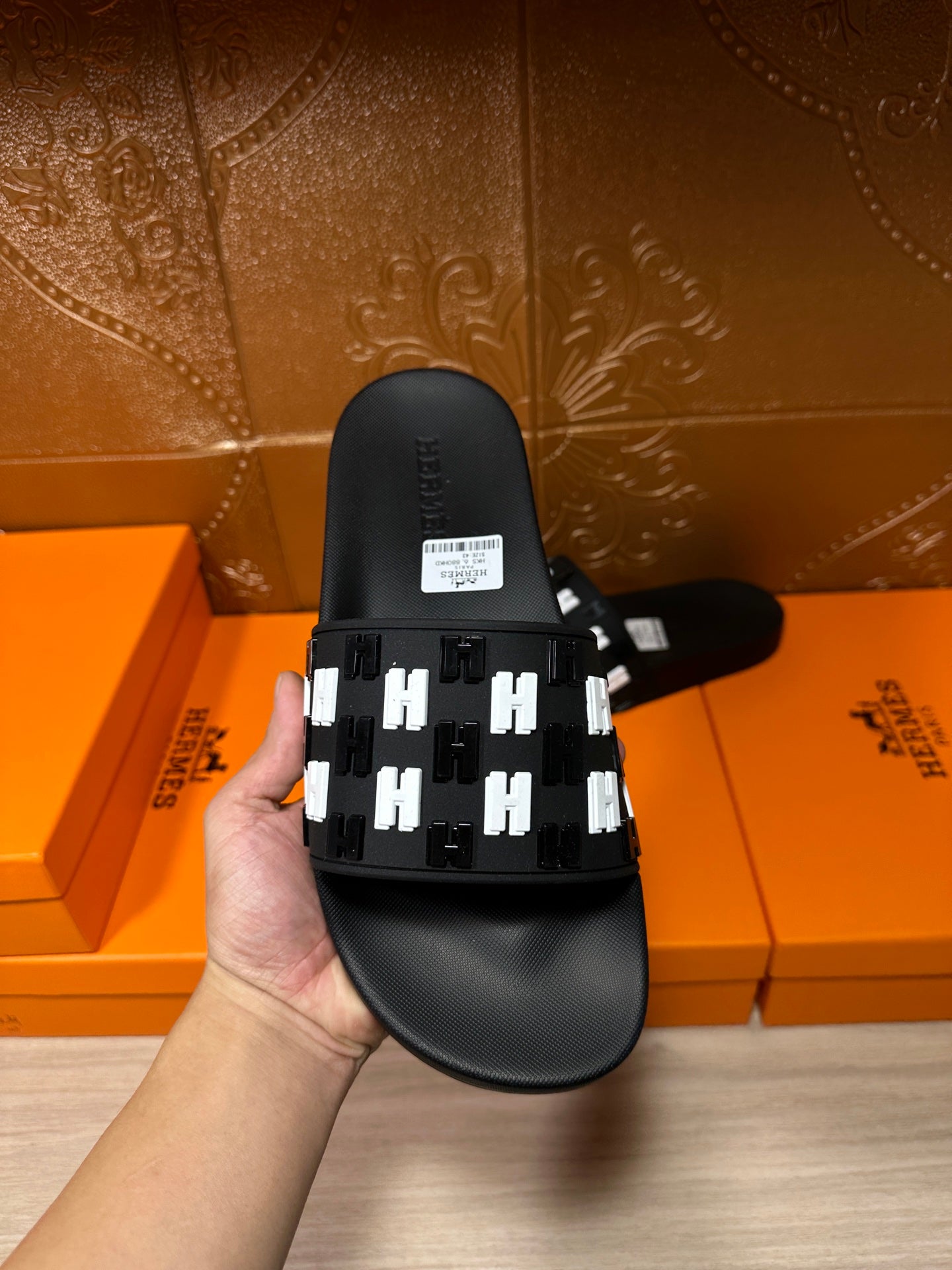 Black slide sandal held up, with white patterned strap and top logo tag