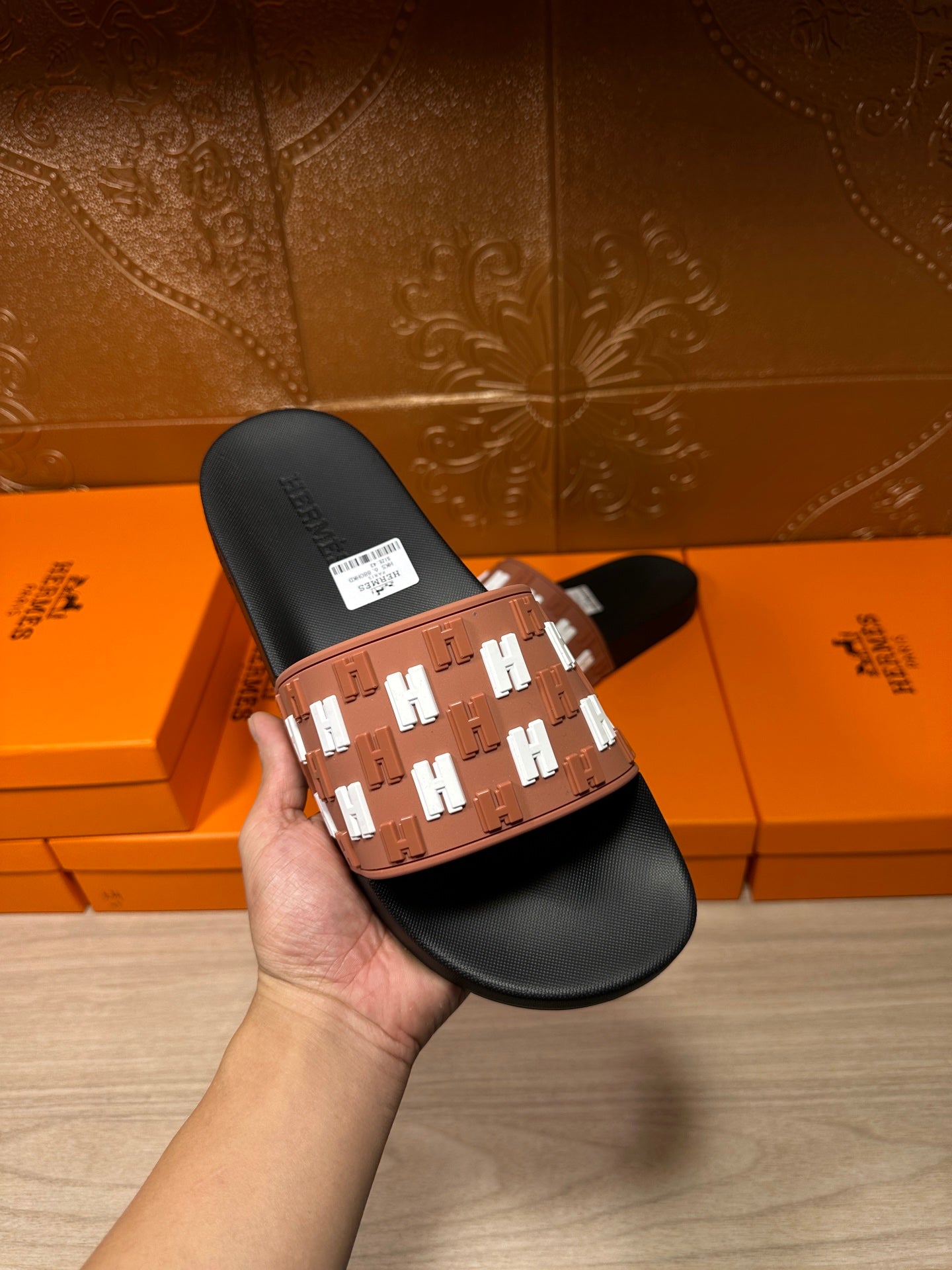 Hermes slide sandal held up, tan strap with white H motifs and black footbed