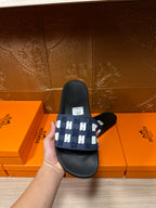 Black slide sandal with navy strap and white H pattern, heel view