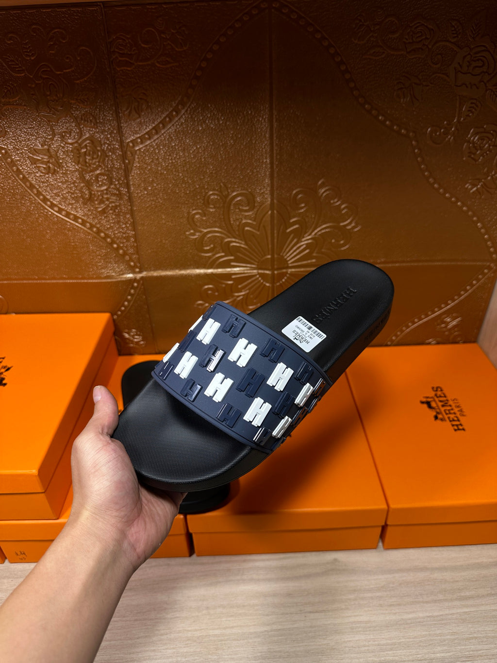Black slide sandal on box; navy strap with repeating white H pattern