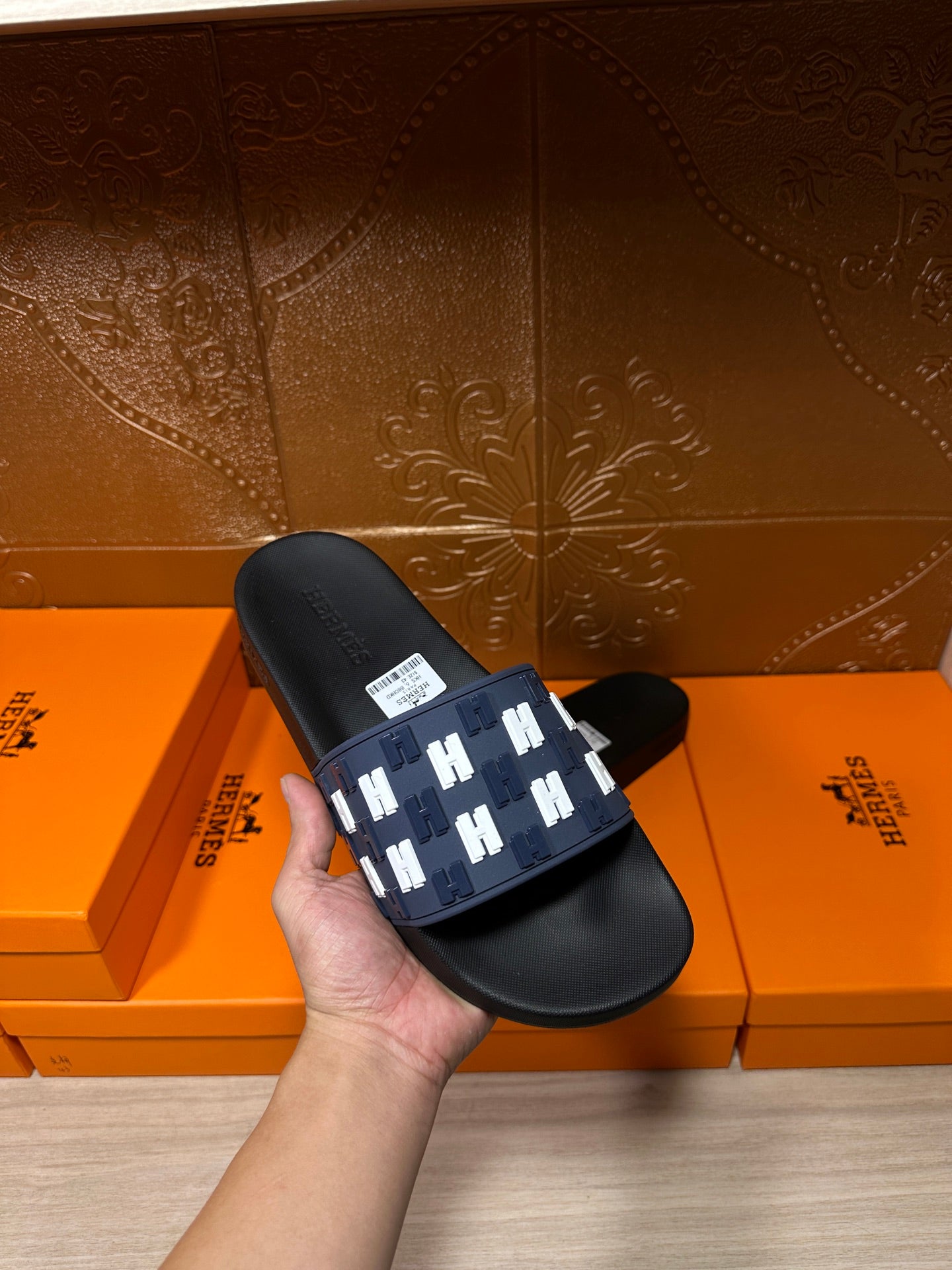 Black slide sandals held in hand; navy strap with white H pattern