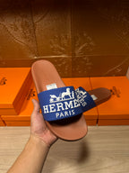 Hand holding brown slide sandal with blue Hermes Paris strap