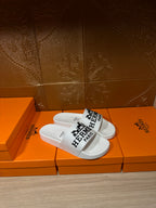 White slide sandals with black Hermes Paris logo across the strap