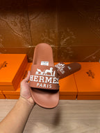 Brown slide sandal held up, white HERMES PARIS logo on strap