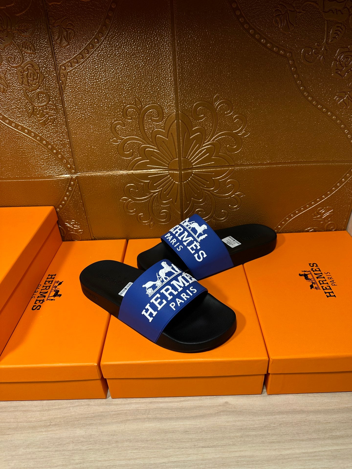 Black slide sandals with blue Hermes Paris logo strap, on orange retail boxes.
