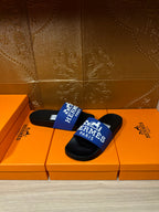Black slide sandals with blue Hermes Paris strap, displayed on orange boxes.