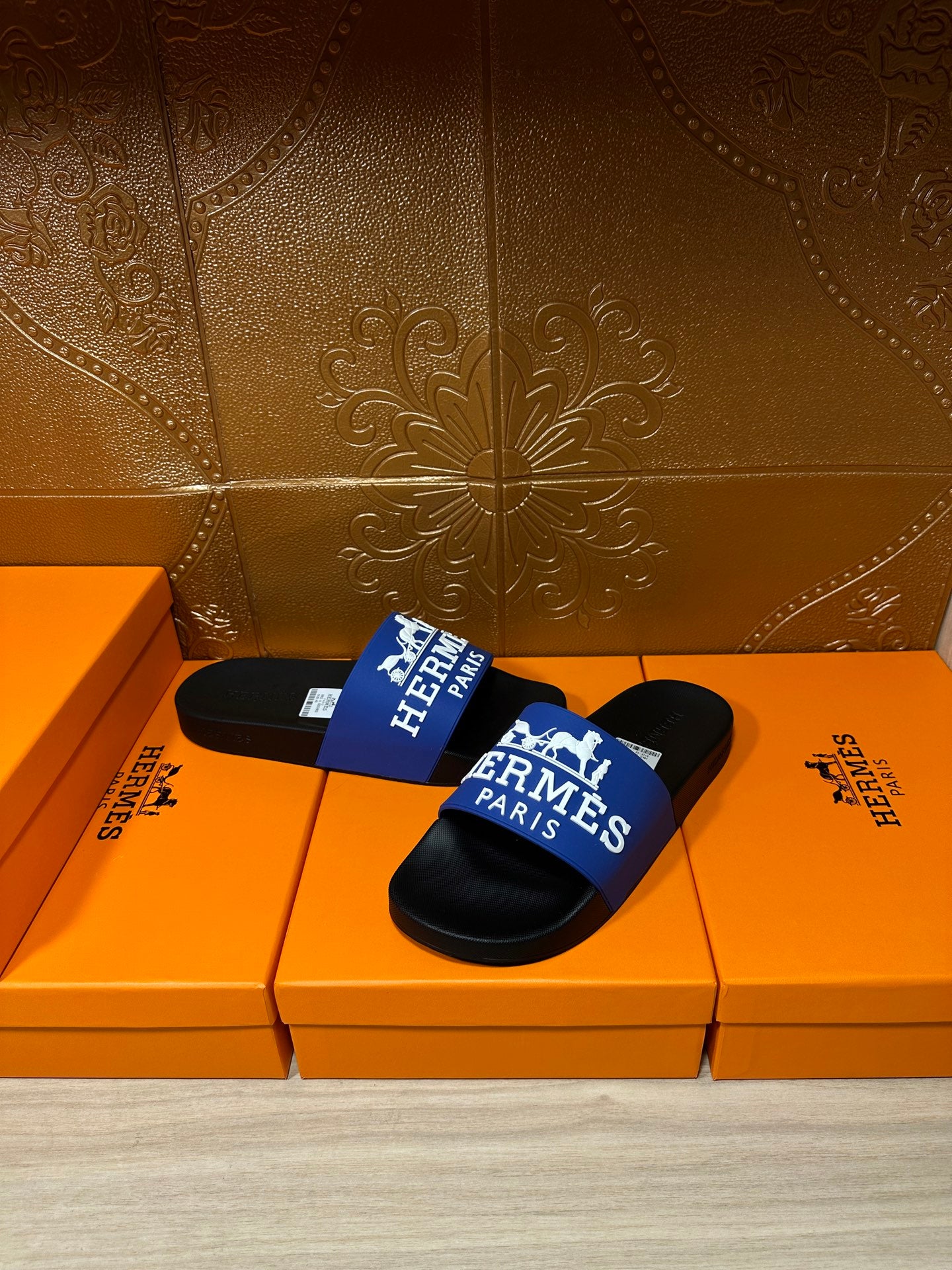 Black slide sandals with blue Hermes Paris strap, displayed on orange boxes.