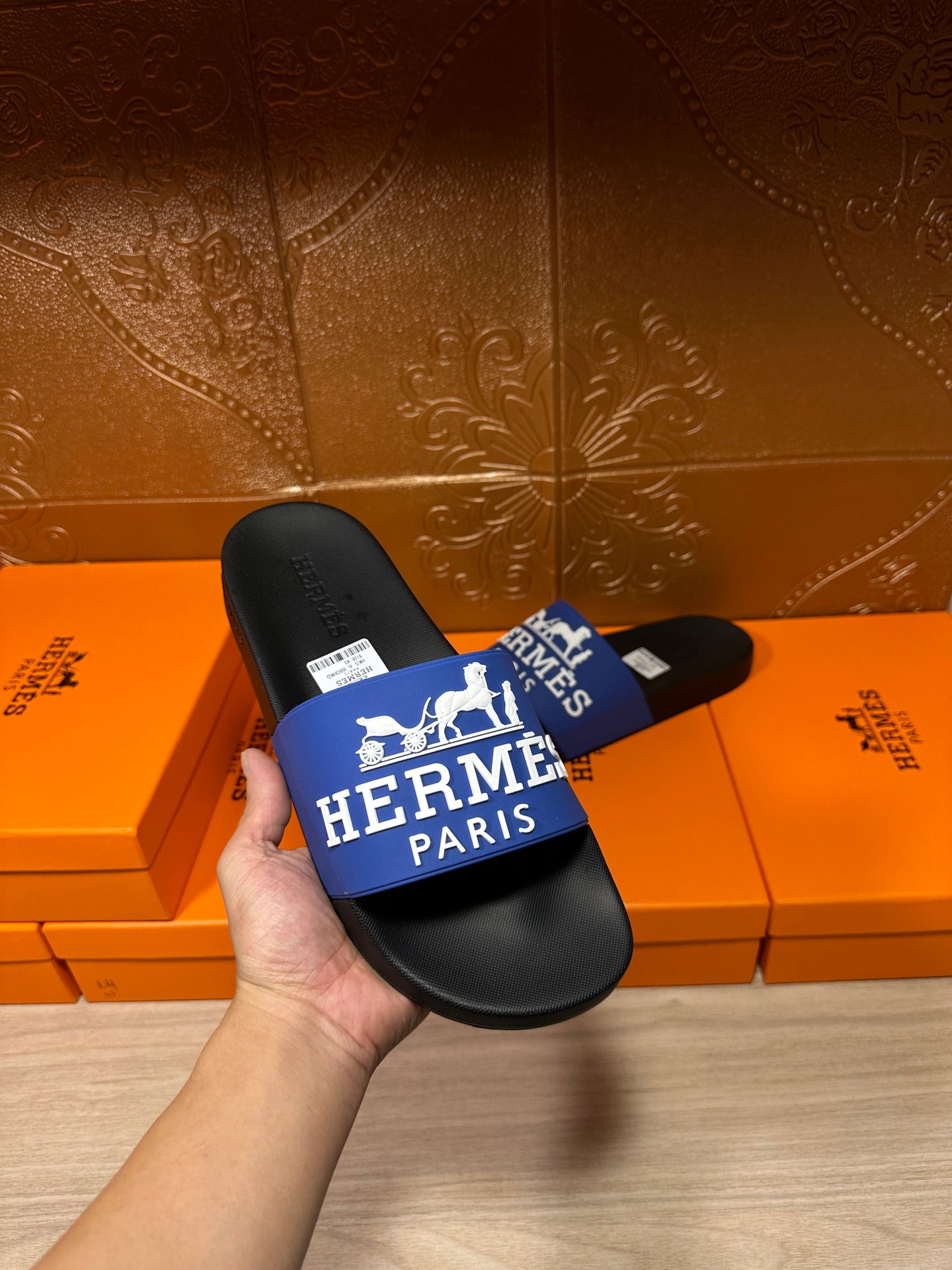Hand holding black slide sandals with blue Hermes Paris strap and white tag.