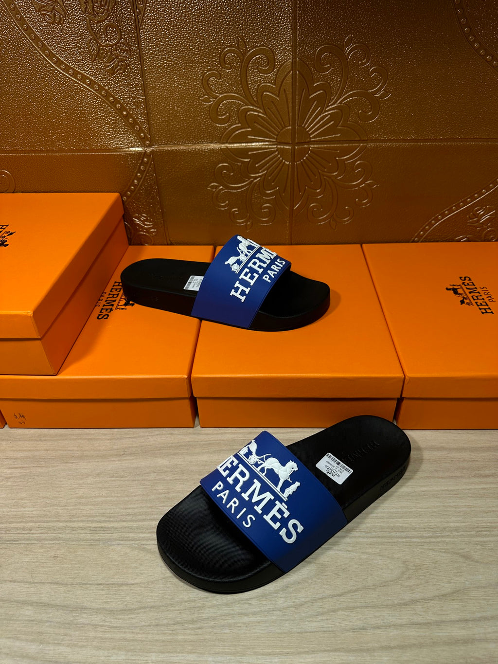 Black slide sandals with blue Hermes Paris logo strap, resting on orange boxes.