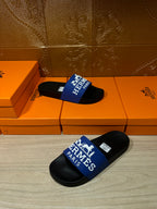 Black slide sandals with blue Hermes Paris logo strap, resting on orange boxes.