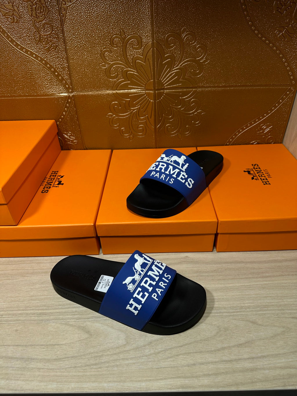Black slide sandals with blue Hermes Paris strap, set on orange retail boxes.