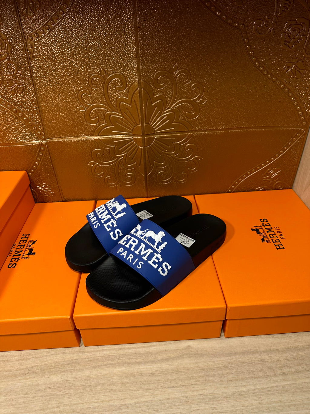 Black slide sandals with blue Hermes Paris logo strap, single pair on orange box.
