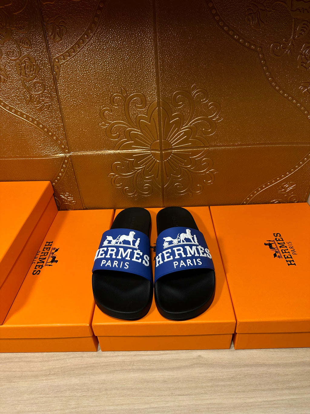 Black slide sandals with blue Hermes Paris strap, shown on orange retail boxes.
