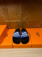 Black slide sandals with blue Hermes Paris strap, shown on orange retail boxes.