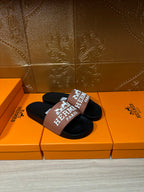 Black slide sandals with tan Hermes Paris logo strap, pair arranged on top view