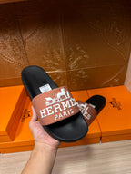 Black slide sandals held in hand with tan Hermes Paris logo strap and tag