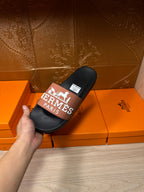 Black Hermes slide sandals held up, tan strap with Hermes Paris logo and tag