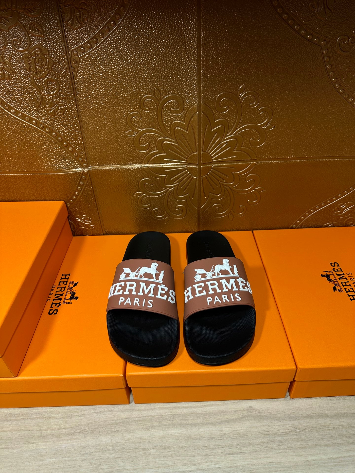 Black slide sandals with tan strap featuring Hermes Paris logo, shown as a pair