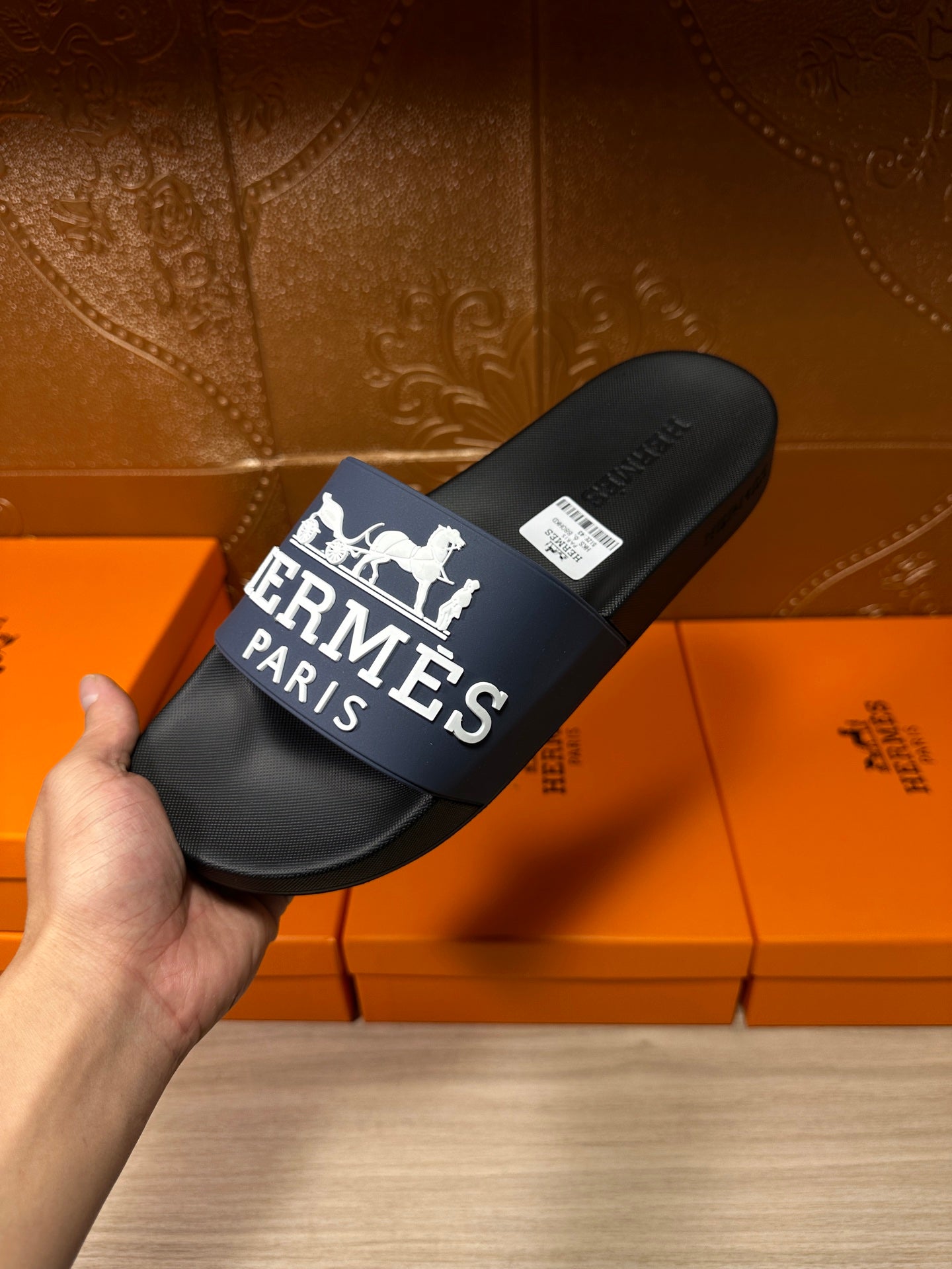 Black slide sandal in hand with blue strap, white horse logo and "Hermès Paris"