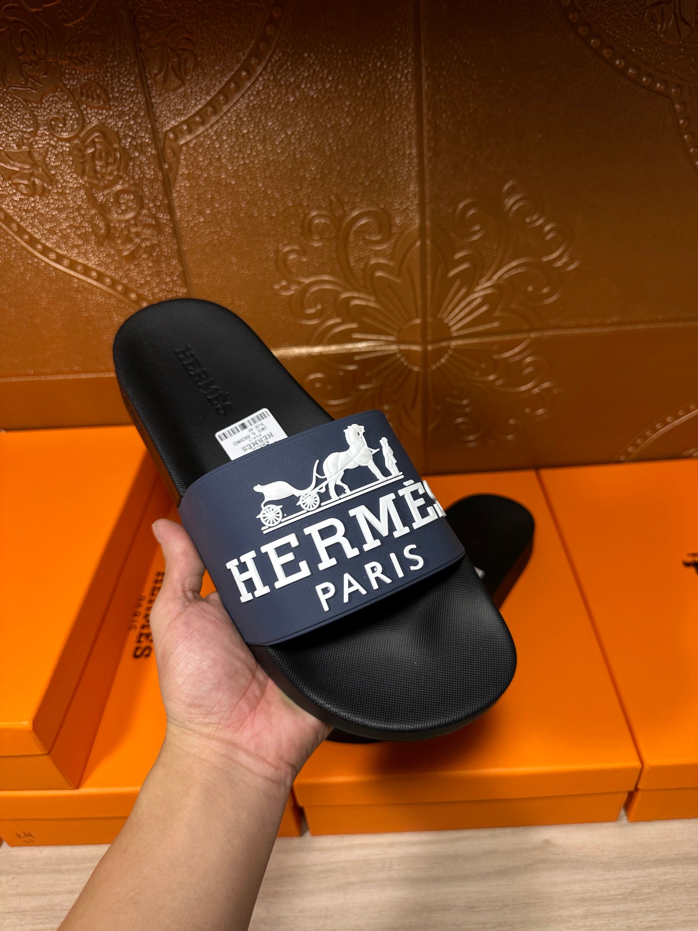 Black slide sandal held in hand with blue strap and white "Hermès Paris"