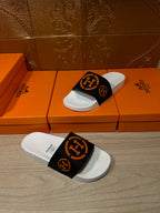 Slide sandal pair: one on the floor, one on a box, black strap with orange logo