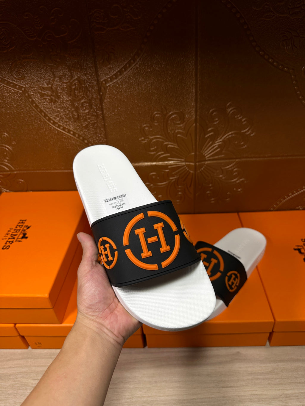 Hand holding Hermes slide sandal: black strap with orange logo, white footbed and sole