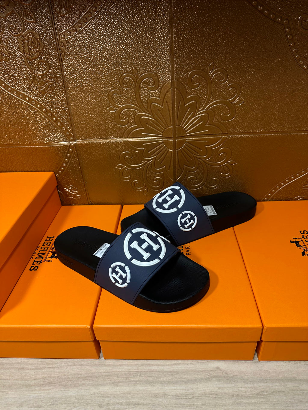 Slide sandals with navy strap and white H logo, black sole, angled on orange box