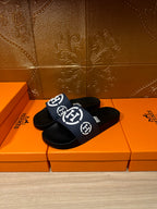 Slide sandals with navy strap and white H logo, black sole, placed on orange box