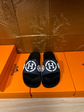 Slide sandals with dark navy strap and white H logo, black footbed, on box