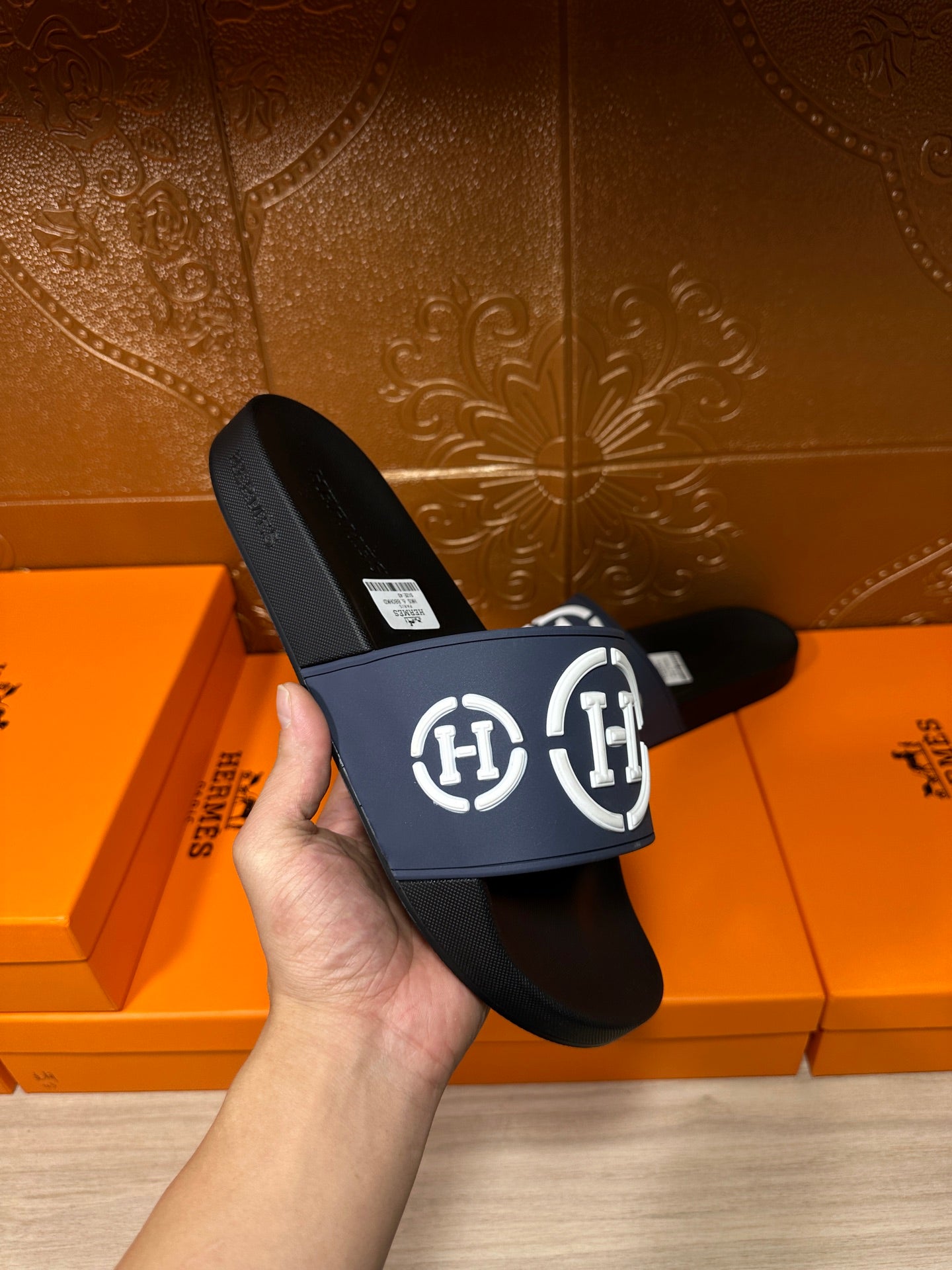 Hand holding slide sandals with navy strap, white H logo, black footbed