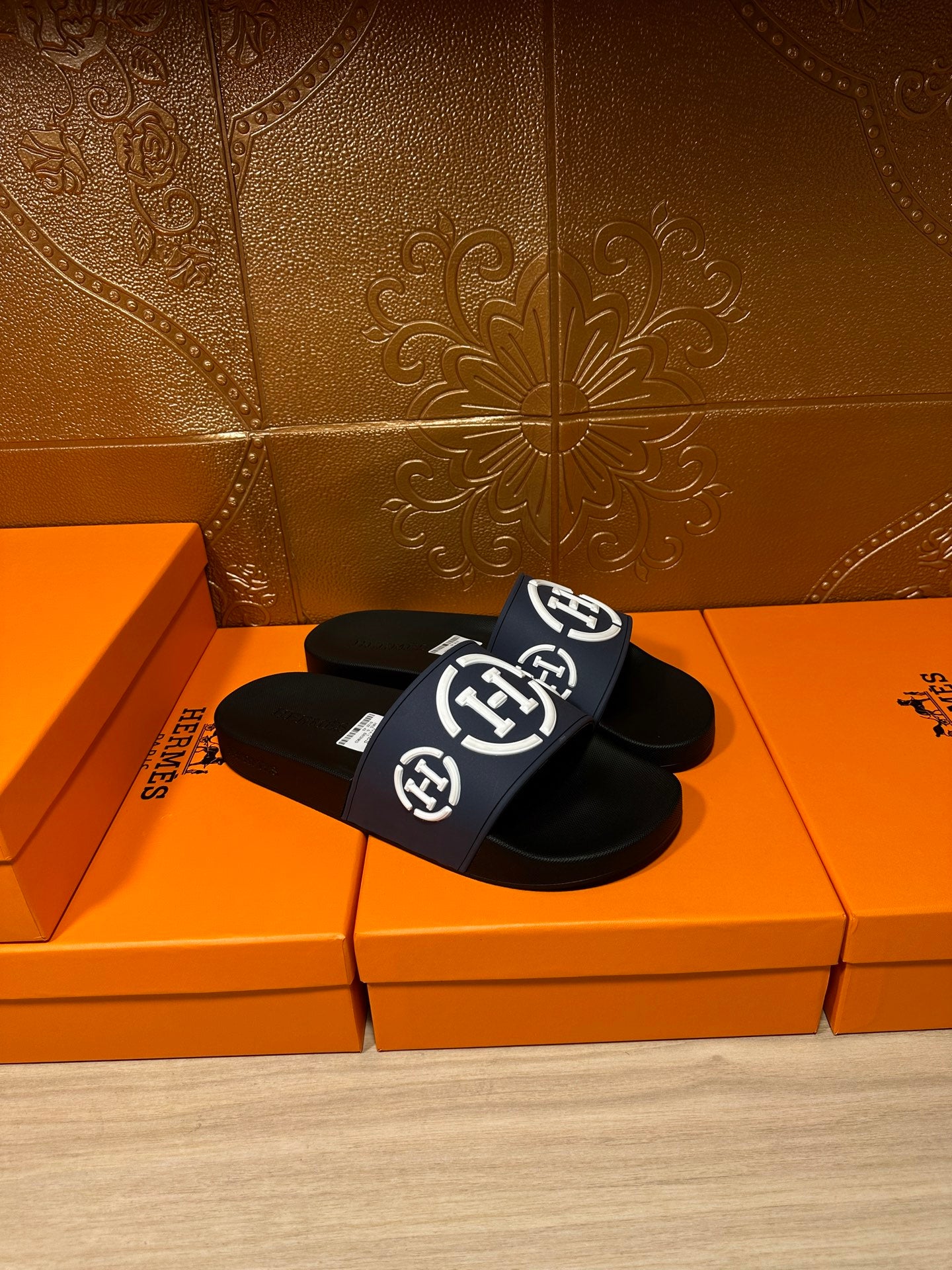 Slide sandals with navy strap and white H emblem, black footbed, on orange box