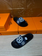 Pair of black slide sandals with navy strap and white H logo, laid on floor