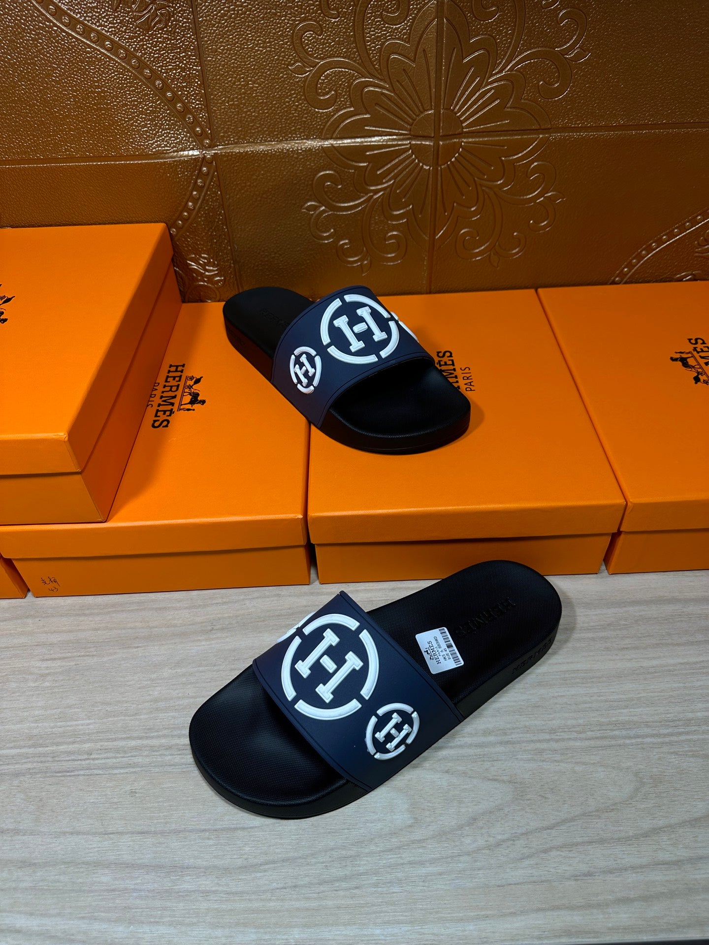 Pair of black slide sandals with navy strap and white H logo, laid on floor