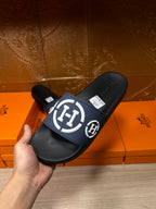 Hand holding slide sandals with navy strap, white H logo, and black footbed