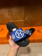 Hand holding black slide sandals with blue logo band and white emblem, flat sole