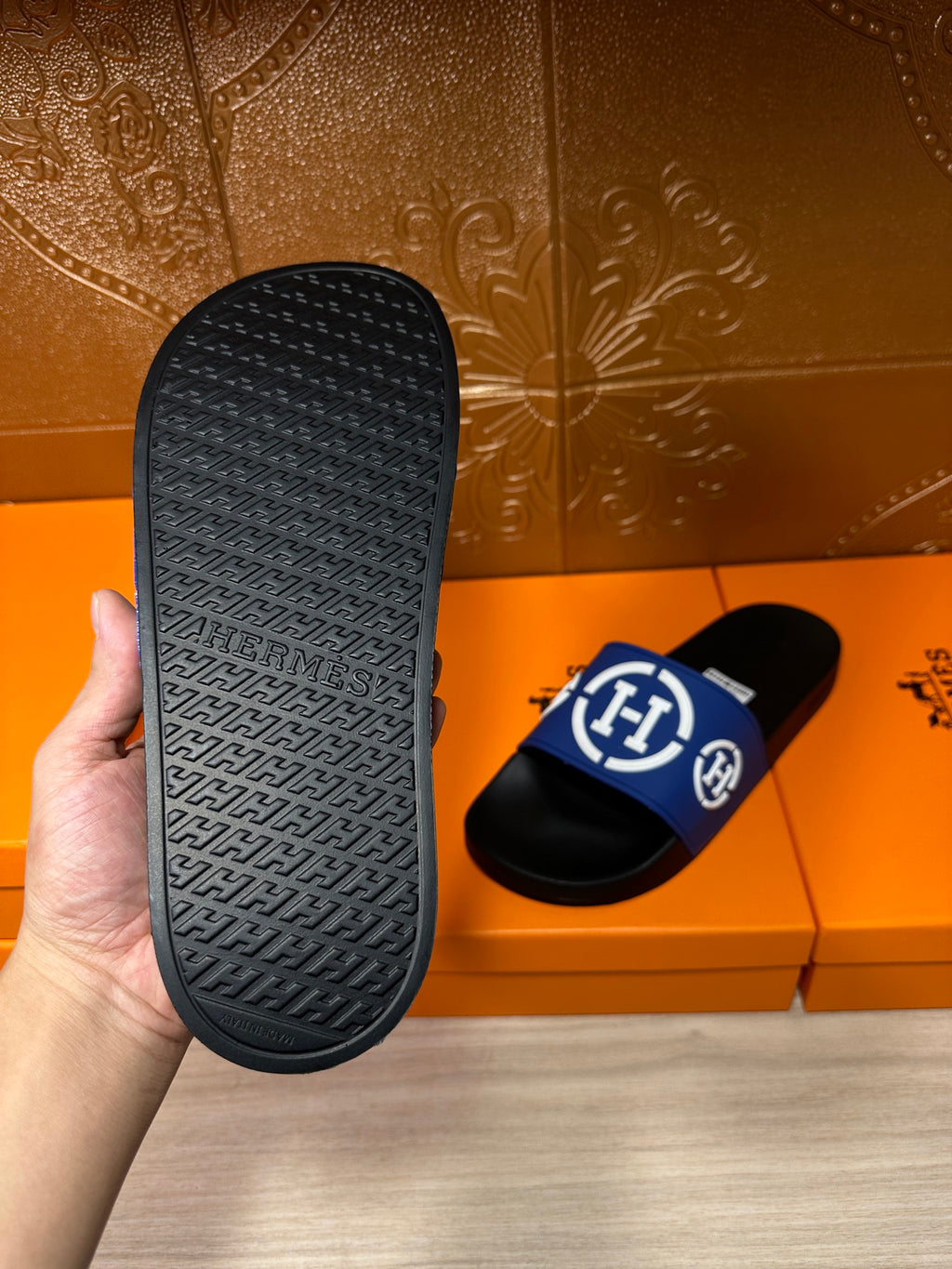 Black rubber sandal outsole with embossed pattern, beside blue logo slide