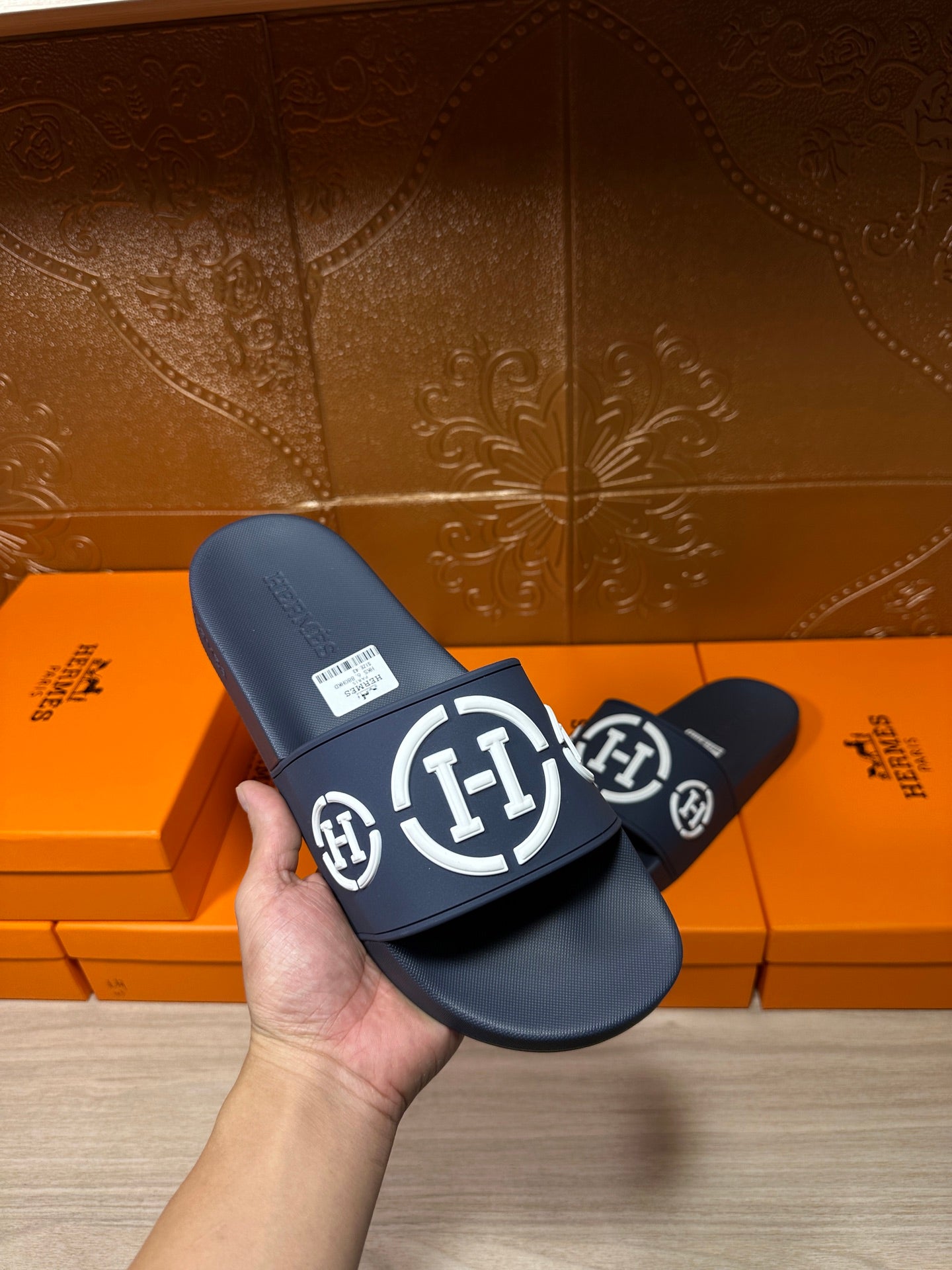 Dark slide sandal held in hand, wide strap with white circular emblem logo