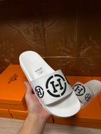 White slide sandals held in hand, black logo strap on top