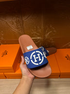 Blue slide sandal with white logo emblem on tan footbed, held in hand