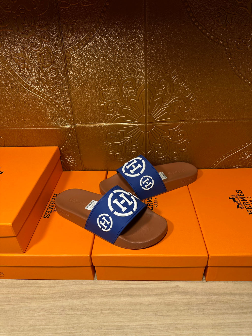 Blue logo slide sandals with brown footbed and small label tag, resting on shoeboxes