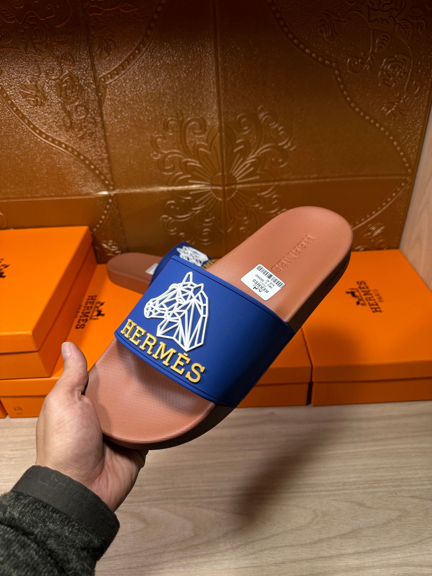 Hand holding Hermes slide sandal with blue logo strap and tan-brown footbed