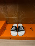 Hermes slide sandals with white footbed and black logo strap, shown on a box display
