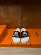 Hermes slide sandals with white footbed and black logo strap, shown on a box display