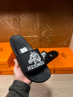 Hermes black slide sandals held in hand, white logo graphic on wide strap