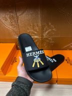 Hermes black slide sandal held in hand, gold logo graphic on strap