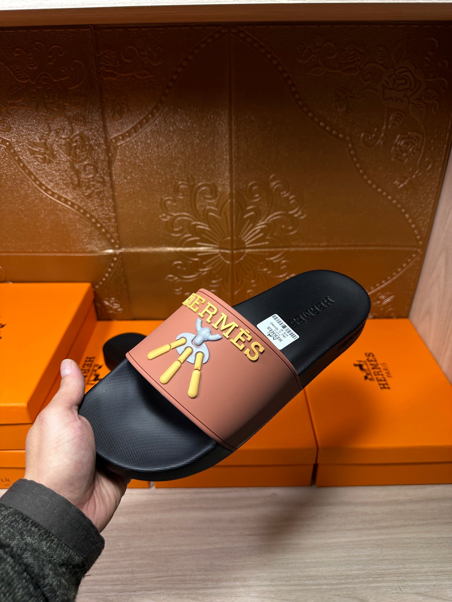 Hand holding Hermes slide sandal: mauve strap, dark footbed, gold Hermes logo and motif.