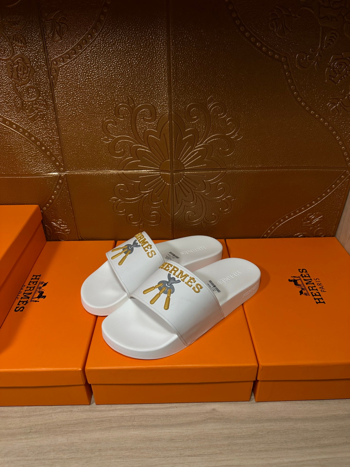 White Hermes slide sandals with gold emblem on the upper, on orange box